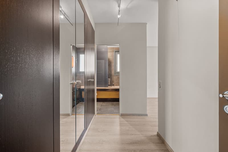 The image shows a hallway with a dark wood-paneled wall on the left, and a doorway leading to a bathroom or kitchen on the right. The floor is a light-colored wood or laminate. There is a dishwasher visible in the adjacent room.