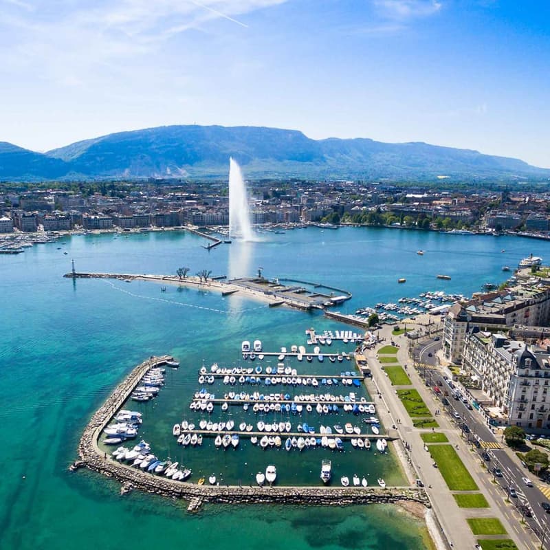 Aerial view of the city of Geneva, Switzerland, with the iconic Jet d'Eau fountain in the center, surrounded by a harbor filled with boats and yachts, and the city's buildings and mountains in the background.