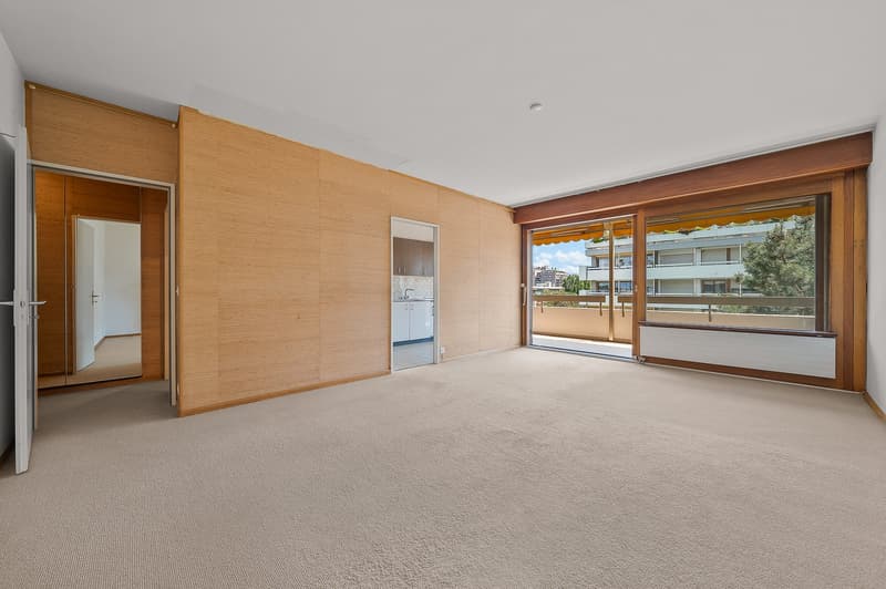 Large open living space with wooden paneled walls, sliding glass doors leading to a balcony, and a carpeted floor.