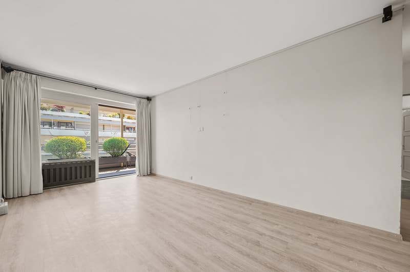 Spacious living room with large windows, hardwood-style flooring, and a balcony or terrace overlooking the outdoor greenery.