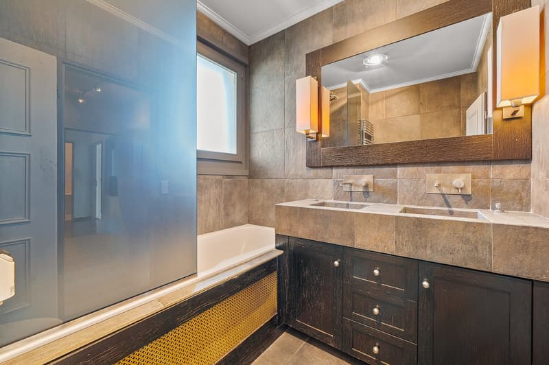 Large bathroom with tiled walls, double vanity with dark wood cabinets, large mirror, and a bathtub