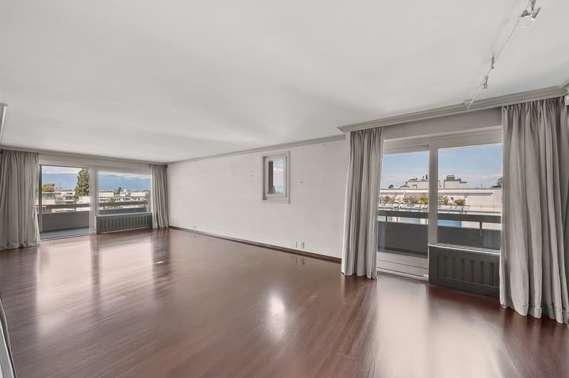 Spacious living room with hardwood floors, large windows, and access to a balcony or terrace. The room has neutral-colored walls and curtains, providing a clean and minimalist aesthetic.