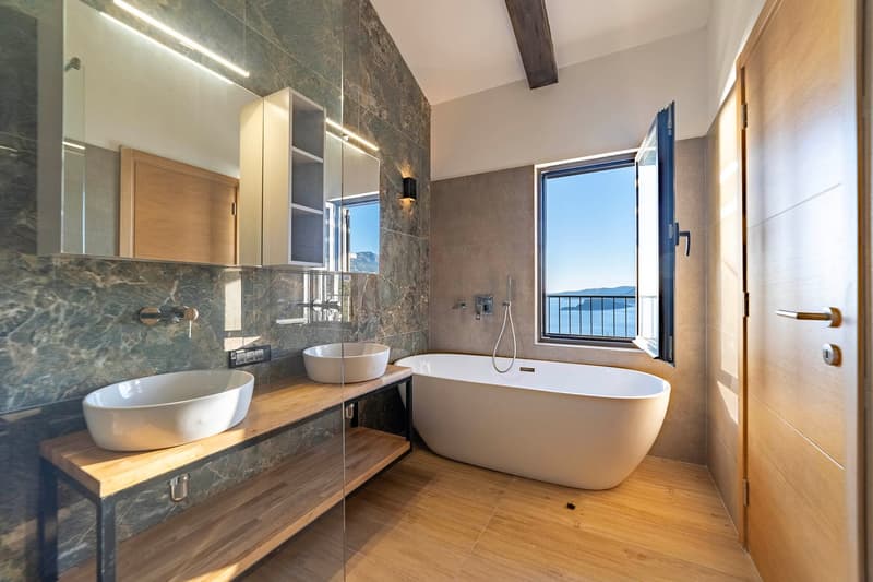 white bathtub, two white sinks, wooden shelf, glass wall, large mirror, wooden door, balcony window