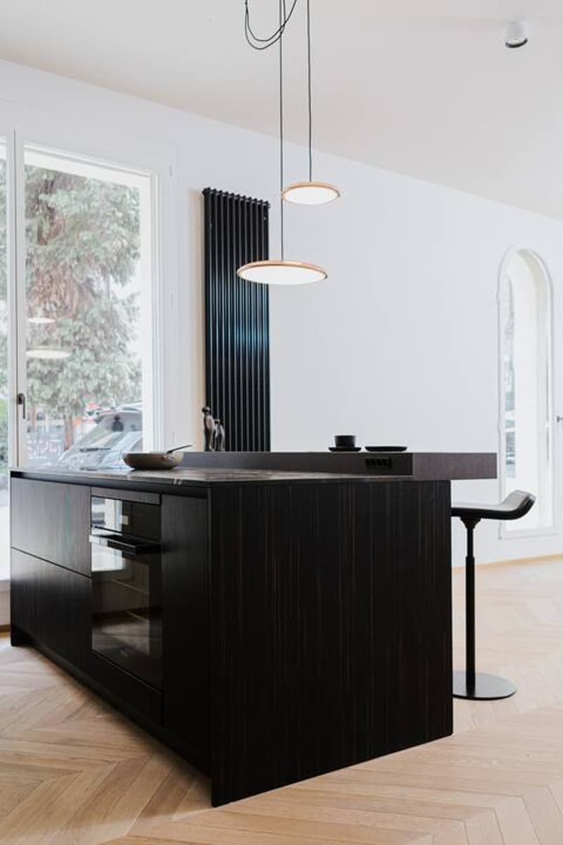 The image shows a modern and minimalist kitchen design. The kitchen features a large black island with a sleek and dark countertop. On the island, there appears to be a dishwasher, which is one of the visible features in the image. The kitchen is well-lit