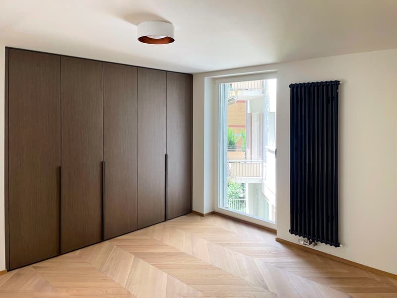Large bedroom with dark brown built-in wardrobes, hardwood herringbone flooring, and a large window with a view of a balcony or terrace.