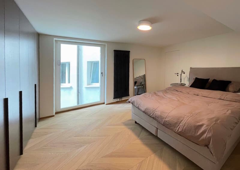 Spacious bedroom with hardwood flooring, large windows, and a sliding glass door leading to a balcony or terrace. The room features a king-size bed, a dresser, and a full-length mirror.