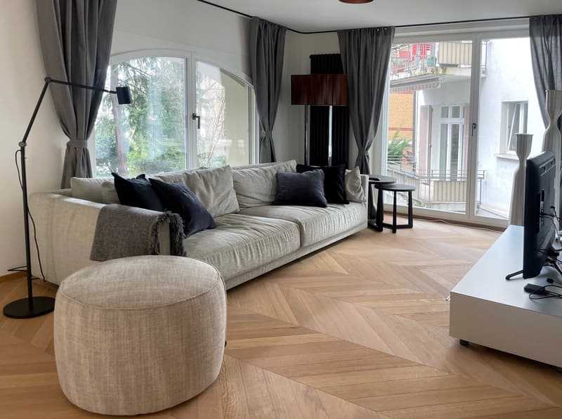 Spacious living room with large arched windows, hardwood herringbone flooring, gray sofa with black accent pillows, black side table, and a floor lamp. The room has access to a balcony or terrace through the windows.