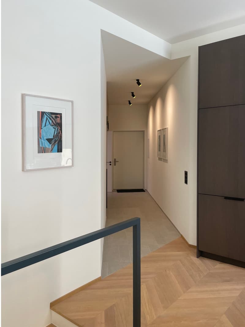 The image shows a hallway with white walls, a framed artwork on the wall, and a wooden floor with a herringbone pattern. The hallway leads to a door and has recessed lighting fixtures on the ceiling.