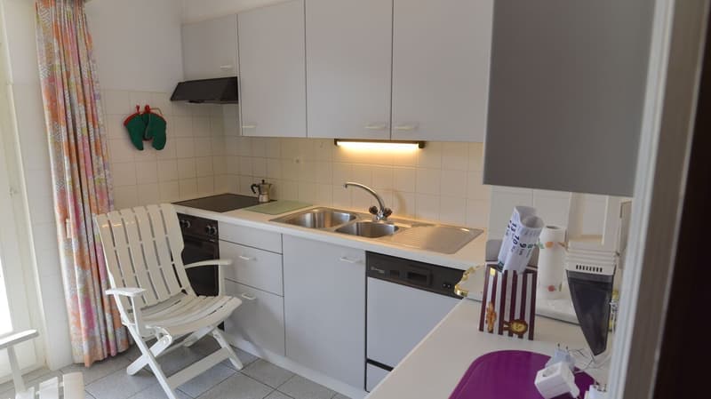 The image shows a small kitchen with white cabinets, a sink, a stove, and a dishwasher. There is a white chair in the corner and some decorative items on the counter.