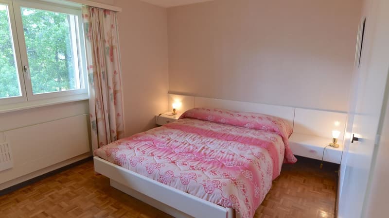 Spacious bedroom with large windows overlooking greenery, white walls, white bed frame with pink floral bedding, wooden flooring, and curtains with floral pattern.