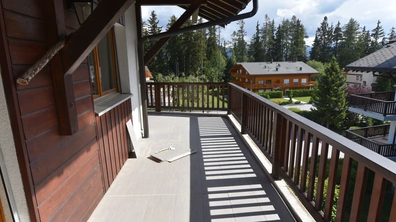 Wooden balcony with railing, overlooking a forested area with cabins in the background