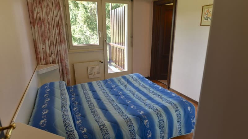 Bedroom with large window overlooking a balcony or terrace, blue patterned bedding, wooden furniture, and a curtained doorway leading to another room.