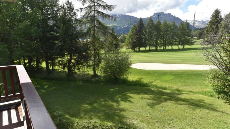 The image shows a scenic golf course surrounded by lush green trees and mountains in the background. The course features a well-manicured fairway and a sand trap. The overall setting appears to be in a picturesque, natural environment.