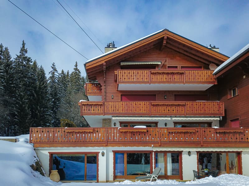 2-story wooden chalet-style house with balconies, surrounded by snow-covered pine trees