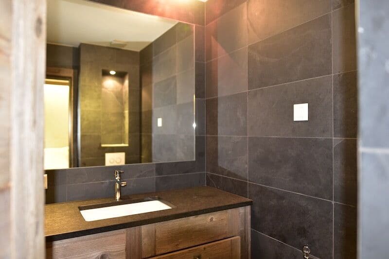 Modern bathroom with dark gray tile walls, a wooden vanity with a sink, and a mirror on the wall.