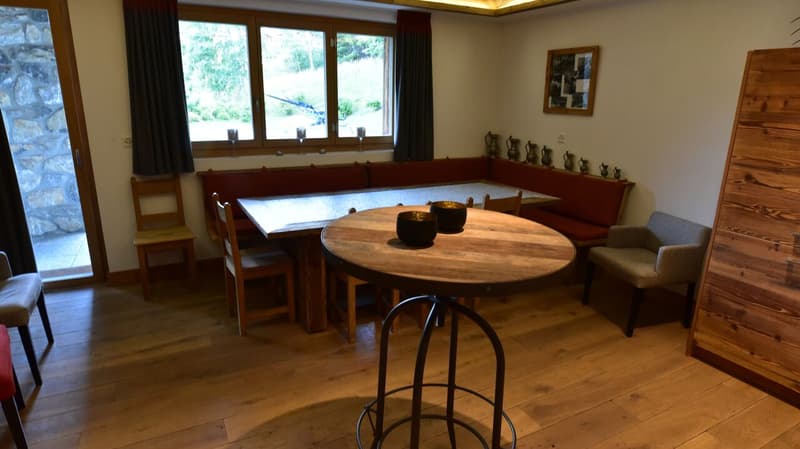 Spacious dining room with large wooden dining table and bench seating, wooden chairs, wooden cabinets displaying decorative vases, and a fireplace. The room has large windows overlooking a scenic outdoor view.