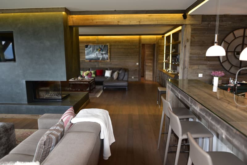 The image shows a spacious open-plan living area with a living room, dining area, and kitchen. The room features wooden beams and paneling, creating a rustic and cozy atmosphere. There is a fireplace visible, and the lighting is warm and ambient. The livi