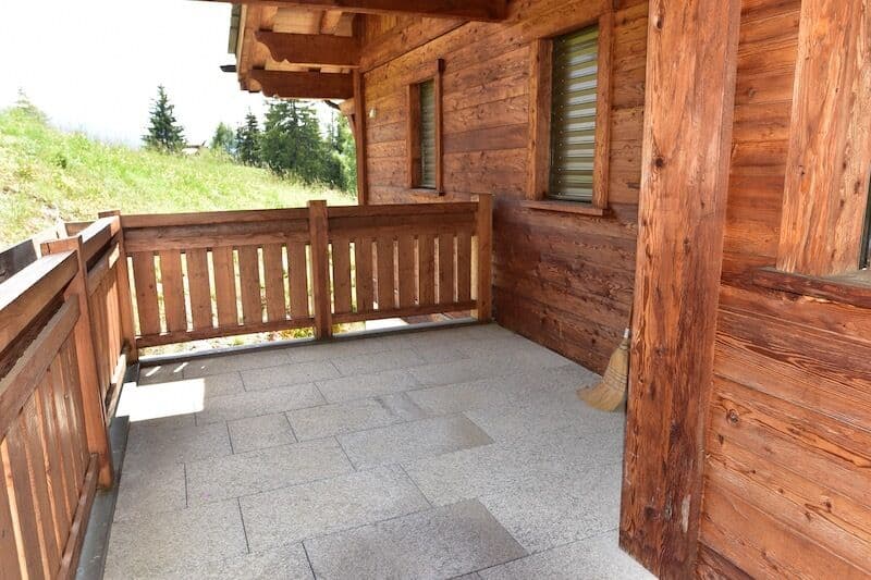 Wooden balcony with railing, stone tile flooring, surrounded by a wooden cabin structure with large windows