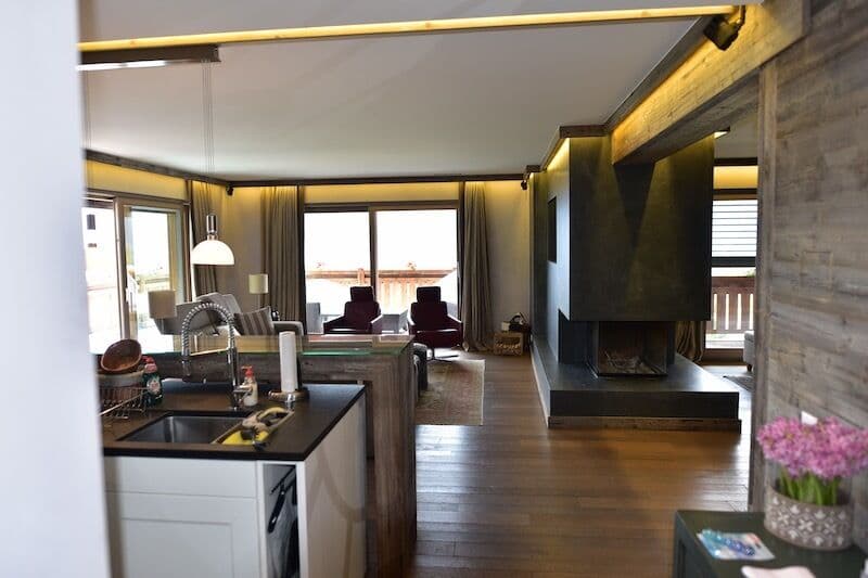 The image shows an open-plan living space with a kitchen area, a living room with a fireplace, and large windows providing a view of the outdoors. The space has a modern, rustic design with wooden floors, stone walls, and warm lighting.