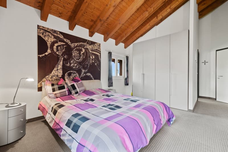 Spacious bedroom with wooden beamed ceiling, large abstract artwork on the wall, queen-sized bed with colorful patterned bedding, built-in wardrobe, and a small side table with a lamp.