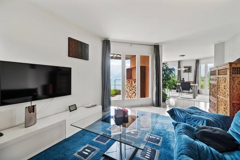 Spacious living room with large windows, glass coffee table, blue sofa, TV, and decorative wall art. The room has access to a balcony or terrace and features a fireplace.