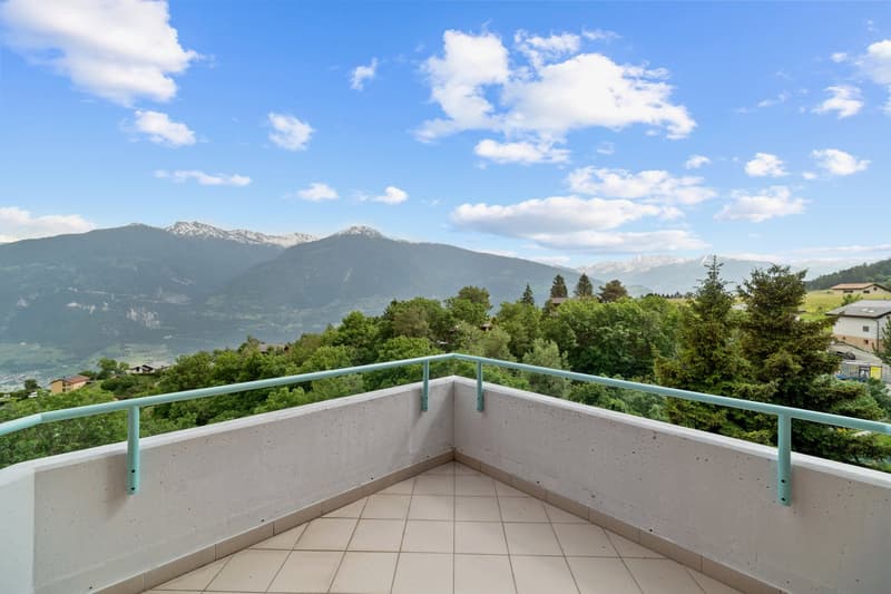 Spacious balcony with a panoramic view of the mountains and lush green trees. The balcony has a concrete floor and a metal railing painted in a light blue color.