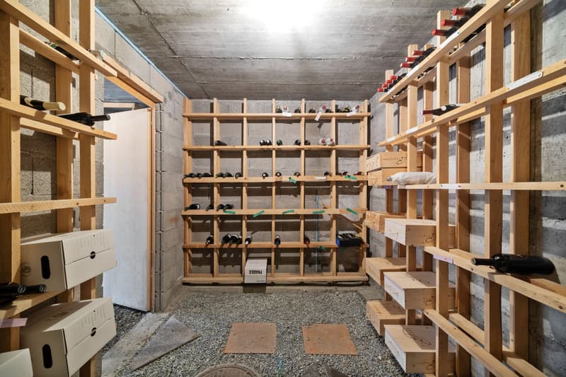 The image shows a large, open space with concrete walls and ceiling. The room is filled with wooden shelving units that are used to store numerous wine bottles. There are also some cardboard boxes and other storage containers visible on the shelves. The f