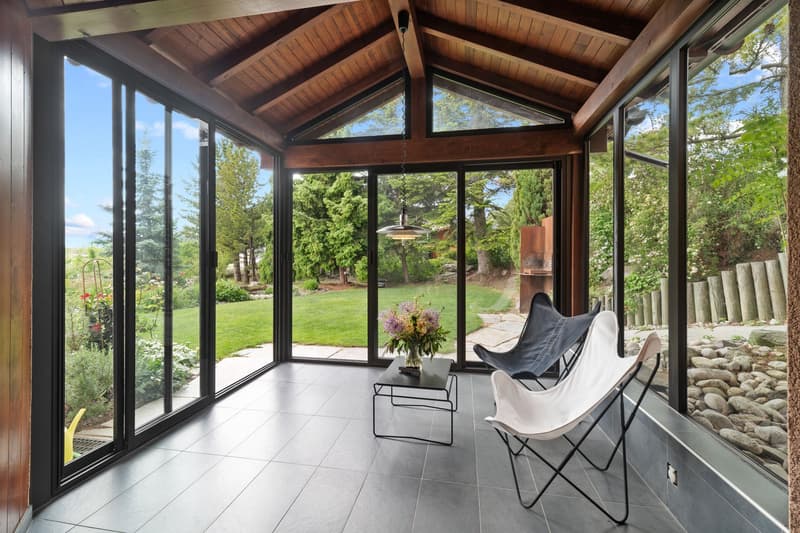 Spacious sunroom with large windows overlooking a lush garden, wooden beamed ceiling, tiled floor, and comfortable seating including butterfly chairs