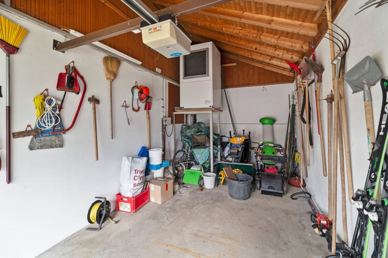The image shows a cluttered garage space with various tools, equipment, and storage items. The garage has a wooden ceiling and walls, and appears to be used for storage and housing various household and gardening items.