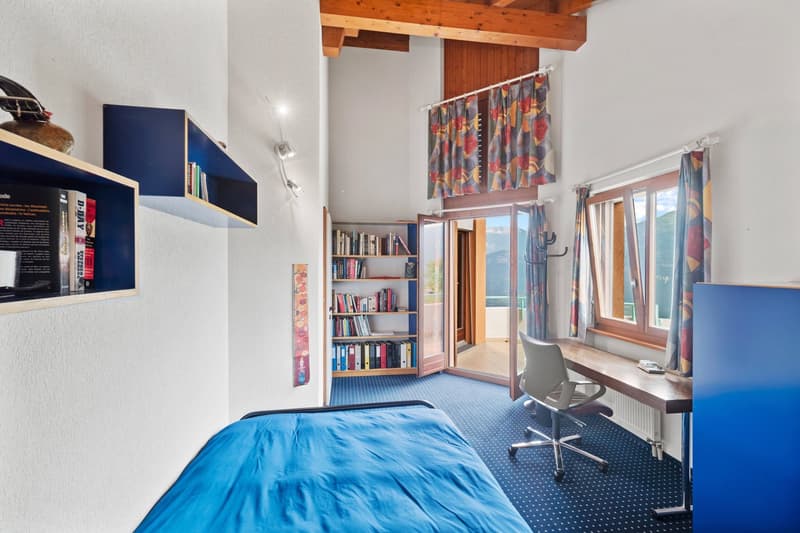 Spacious bedroom with wooden ceiling beams, large windows providing natural light, blue and white color scheme, bookshelf, desk, and chair