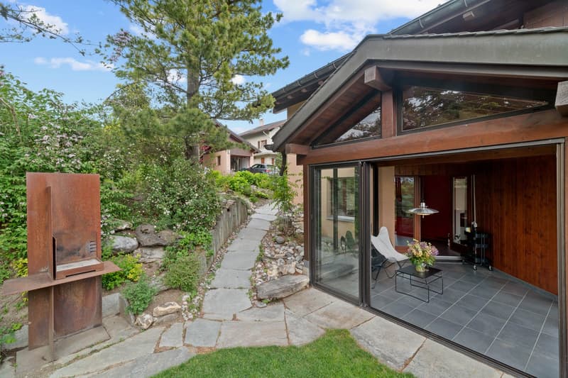 Two-story wooden house with a covered patio, large windows, and a stone pathway leading to the entrance. The house is surrounded by lush greenery, including pine trees and other vegetation. There is a stone fireplace visible on the exterior of the house.