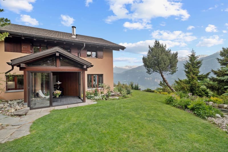 2-story wooden house with a balcony, surrounded by a well-maintained garden and lawn, with a scenic mountain view in the background