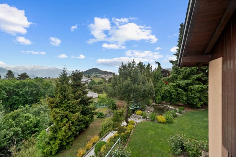 The image shows a scenic view of a residential area surrounded by lush greenery and mountains. The house appears to be situated on a hillside, with a well-maintained garden and landscaping visible in the foreground. The wooden structure of the house blend