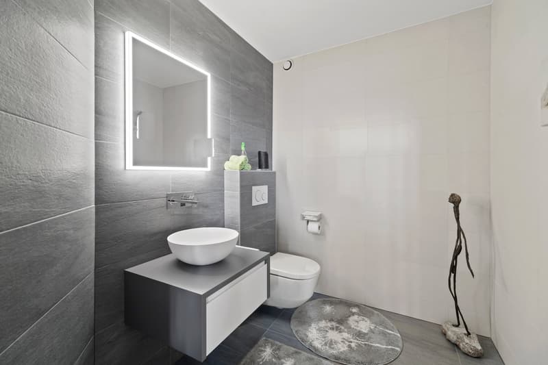 Modern bathroom with gray tile walls, white vanity with vessel sink, backlit mirror, and minimalist decor elements like the sculpture and rug.