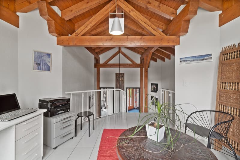 Spacious open-concept living area with high vaulted wooden ceiling, exposed wooden beams, and a large window. The room features a desk with a computer, a printer, and other office equipment, as well as a seating area with a chair and a plant. The space al