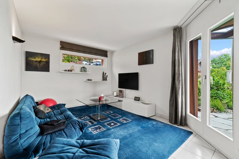 Spacious living room with large windows, blue velvet sofa, glass coffee table, wall-mounted TV, shelves, and access to a balcony or terrace.