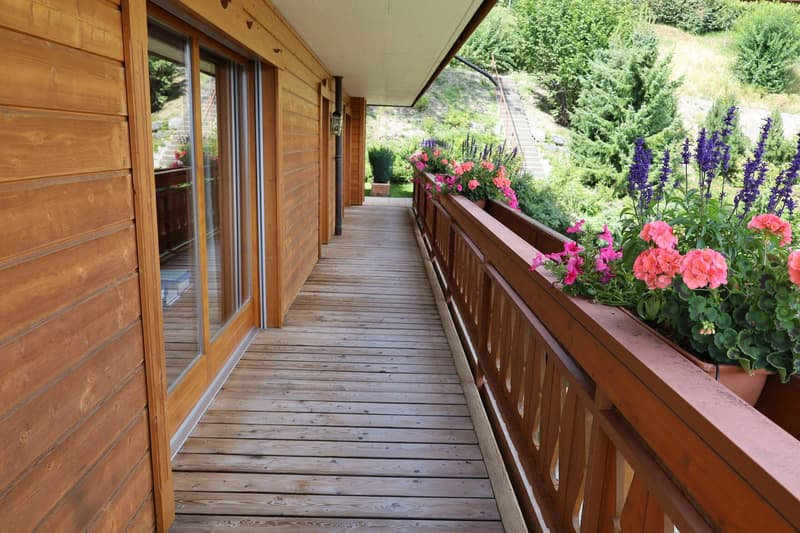 Wooden balcony with sliding glass doors, overlooking a lush garden with colorful flowers and greenery