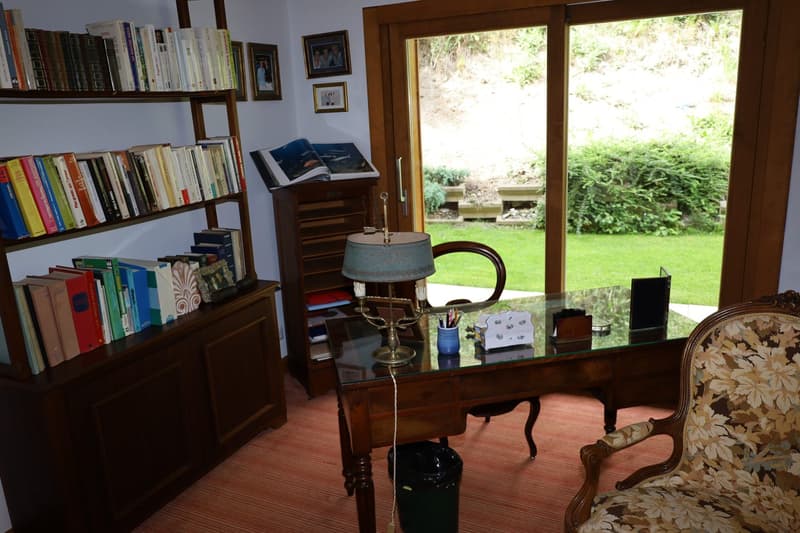 Cozy home office with wooden bookshelves, a desk with a lamp, and a large window overlooking a lush garden. The room has a warm, inviting atmosphere with personal touches like framed artwork and decorative items.