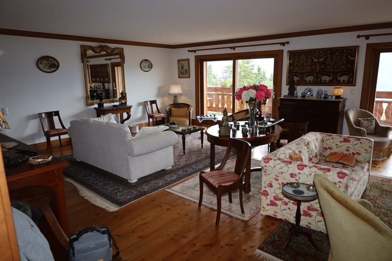 Spacious living room with hardwood floors, white walls, and large windows overlooking a balcony or terrace. The room is furnished with a sofa, armchairs, a coffee table, and a dresser. Decorative elements include framed artwork, mirrors, and floral arrang