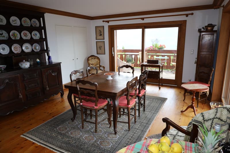 Spacious dining room with wooden furniture, including a large dining table and chairs with pink cushions. The room features a built-in shelving unit displaying decorative plates. Large windows provide access to a balcony or terrace, allowing natural light
