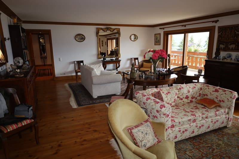 Spacious living room with hardwood floors, white walls, and wooden beams. The room features a floral patterned sofa, a round wooden coffee table, and a large mirror on the wall. There is also a balcony visible through the window, providing natural light a