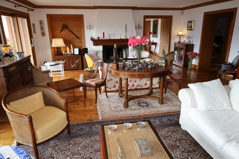 Spacious living room with wooden beams, fireplace, antique furniture, floral patterned chairs, round wooden dining table, and various decorative items and artwork on display.
