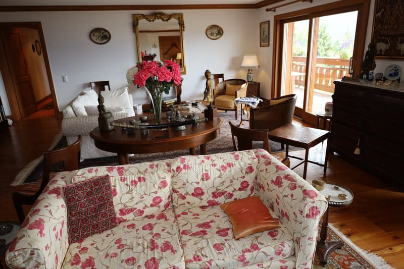 Cozy living room with wooden furniture, floral patterned sofa, ornate mirror, and a vase of pink flowers on a round wooden table. The room has large windows overlooking a balcony or terrace.
