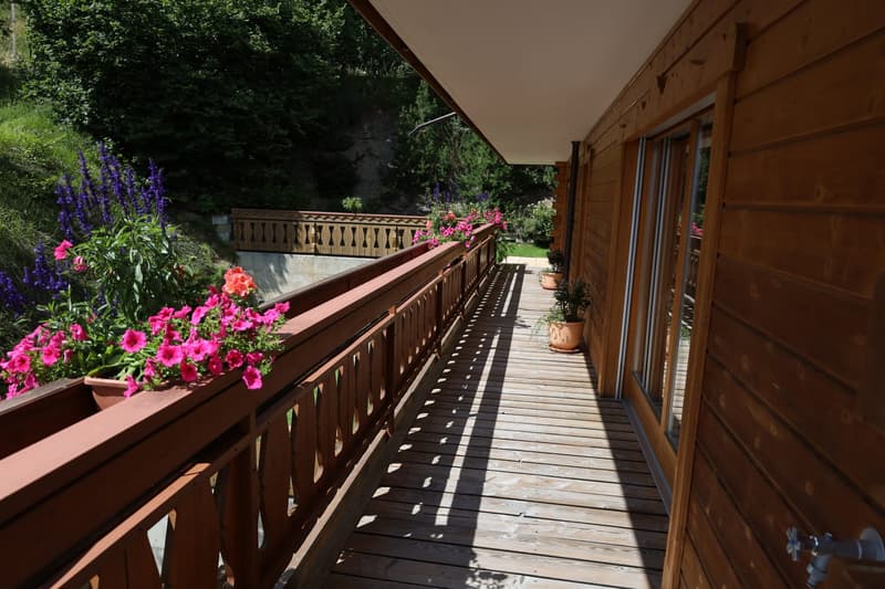 Wooden deck with a railing, surrounded by lush greenery and colorful flowers. The deck leads to a wooden cabin or structure with large windows.