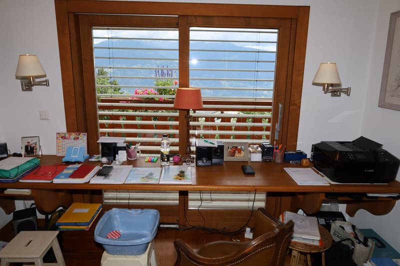Cluttered home office with a large wooden desk, various office supplies and electronics, and a scenic view through the window overlooking a mountainous landscape.