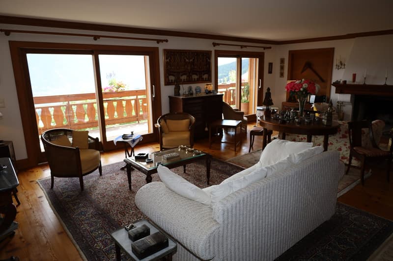 Spacious living room with large windows overlooking a balcony, wooden beams on the ceiling, a cozy sofa, armchairs, a coffee table, and a dining area with a round table and chairs. The room is decorated with various ornaments and artwork on the walls.