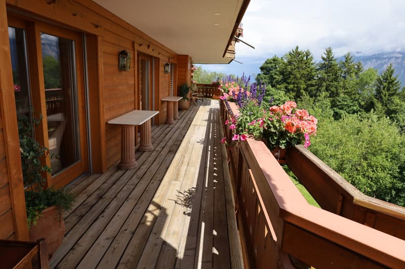 Wooden cabin with a large balcony/terrace overlooking a forested area. The balcony has wooden railings and columns, and is decorated with potted flowers. The cabin appears to be surrounded by trees and greenery.