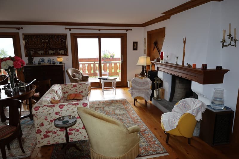Cozy living room with hardwood floors, wooden beams on the ceiling, large windows overlooking a balcony, floral patterned sofa, armchair, and rug, fireplace with a mantel, and various decorative items and artwork on the walls and shelves.