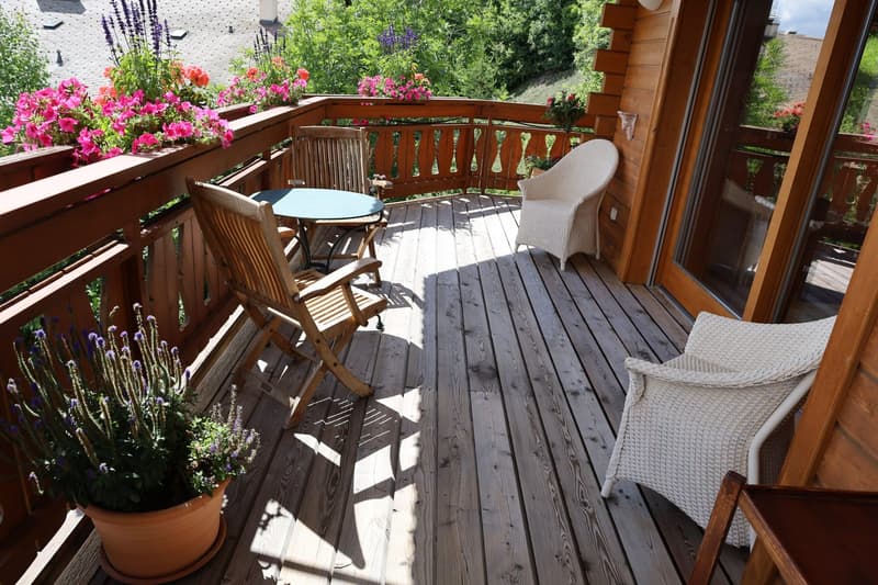 Wooden balcony with a table, chairs, and potted plants. The balcony overlooks a lush, green garden with colorful flowers.