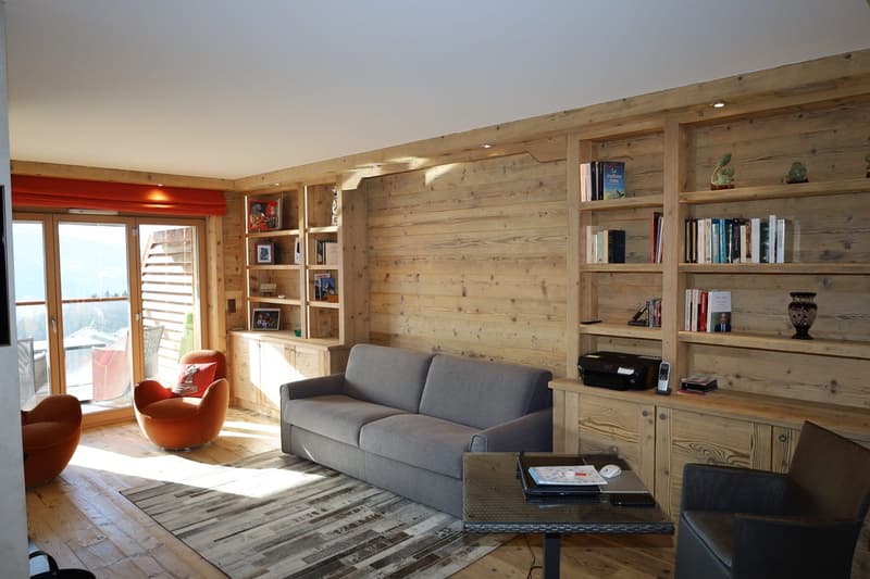 living room with wooden walls, couch, orange armchair, glass door, glass windows, wooden cabinet with shelves, wooden desk with laptop, chair
